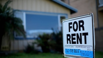 Un cartel blanco y azul de 'FOR RENT' con un número de teléfono y un precio de $1600 visible, ubicado frente a una casa residencial desenfocada