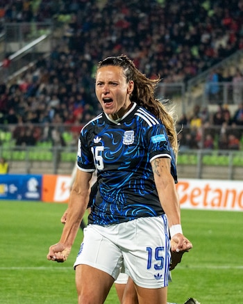 Once jugadoras de la Selección Femenina de Argentina posan sonrientes en el campo de un estadio por la noche, vistiendo uniformes oscuros con detalles celestes y blancos