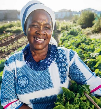 Primer plano de una mujer mayor africana sonriendo, con un turbante y vestido blanco y azul, sosteniendo verduras verdes