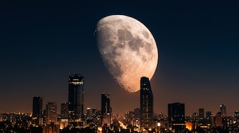 Cielo nocturno con una luna en fase menguante sobre una hilera de rascacielos y luces urbanas.