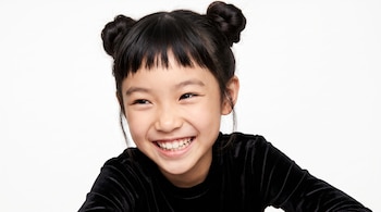Niña sonriente con flequillo y dos moños altos que viste una prenda negra frente a un fondo blanco.