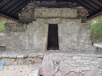 Edificio de piedra con paredes de bloques irregulares, entrada rectangular oscura, techo de paja y vegetación al fondo