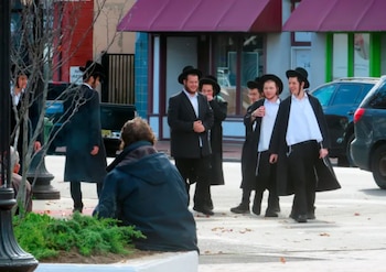 Varios hombres con sombreros negros y abrigos largos caminan por una acera de la ciudad, con un hombre sentado de espaldas en primer plano y coches aparcados al fondo