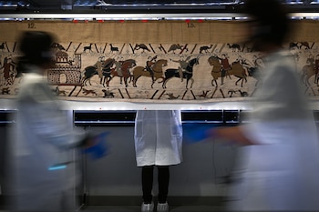 Un gran tapiz antiguo bordado con figuras de personas a caballo, animales y texto en latín. Tres figuras humanas borrosas con batas blancas
