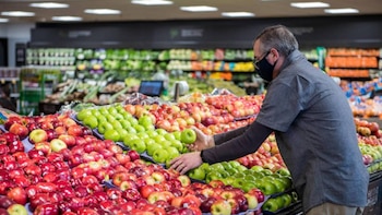Um homem de meia-idade usando máscara preta e óculos escolhe uma maçã verde em uma grande vitrine de frutas vermelhas e verdes em um supermercado.