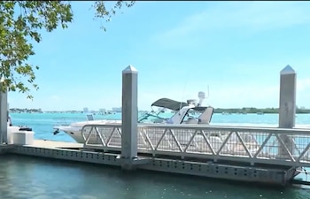 Vista de un muelle con barcos amarrados en aguas turquesas bajo un cielo despejado. Árboles verdes se ven a la izquierda, y edificios en la distancia