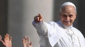 Primer plano de un hombre calvo sonriente con un solideo blanco y vestiduras clericales, señalando con su mano derecha. Otras manos se ven al fondo
