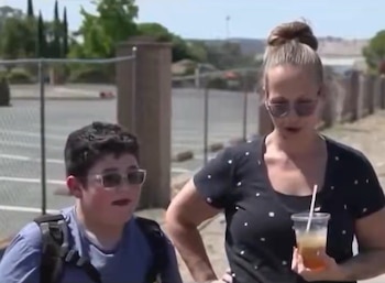Un niño con mochila y gafas junto a una mujer con el cabello recogido y gafas de sol que sostiene un vaso con bebida