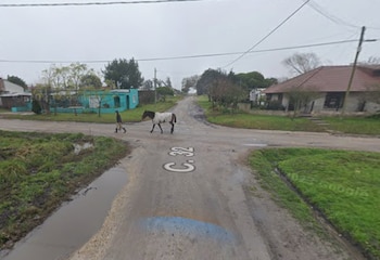 Cruce de caminos de tierra con charcos, casas bajas, árboles y una persona que cruza a pie junto a un caballo cubierto con una manta