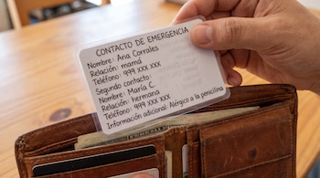 Mano sosteniendo una tarjeta plastificada con datos de emergencia sobre una billetera de cuero marrón.