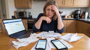 Mujer adulta sentada a una mesa de madera con laptop, recibos de papel, dos teléfonos móviles con pantallas de pago, y sobres en una cocina.