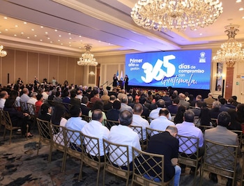 Vista general de un gran salón de conferencias con una audiencia sentada frente a un escenario. Una oradora y una pantalla gigante con texto "365 días de compromiso" son visibles