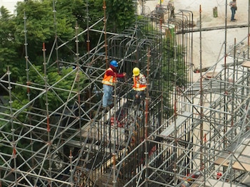 Vista aérea de una obra en construcción con andamios metálicos, dos trabajadores con cascos y chalecos de seguridad en una estructura de varillas de acero