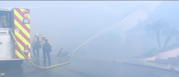 Tres bomberos y la parte trasera de un camión de bomberos en una calle cubierta de humo mientras lanzan agua con una manguera
