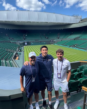 Tres hombres, Mariano Puerta, Brandon Nakashima y un tercero, posan en un estadio de tenis vacío de Wimbledon con asientos verdes y un techo retráctil visible
