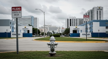 Boca de agua con candado en primer plano, con señales rojas de 'Peligro: Corte de Agua' frente a edificios de condominios y hospitales de Hialeah bajo cielo nublado.