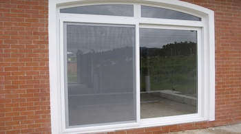 Ventana de marco blanco con mosquitero y vidrio transparente, sobre una pared de ladrillo rojo. Al fondo se observa vegetación y cielo nublado.