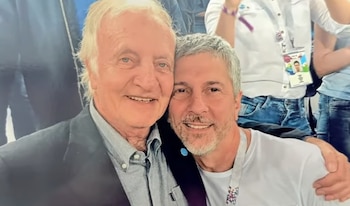 Primer plano de dos hombres sonrientes abrazándose. El hombre de la izquierda tiene cabello blanco y viste chaqueta oscura. El de la derecha tiene barba gris y camiseta blanca