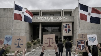 Edificio del Tribunal Constitucional de República Dominicana con banderas nacionales. Manos sostienen carteles con balanzas de justicia, corazones y el escudo nacional.