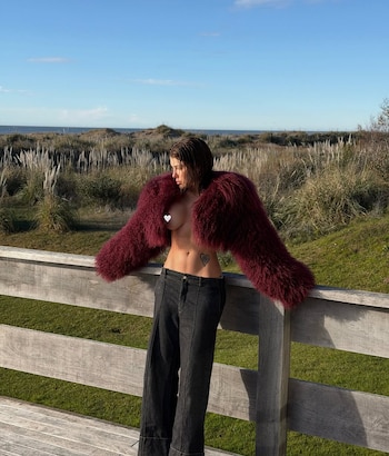 Una mujer de pelo corto y torso desnudo usa una chaqueta de pelo roja y pantalones oscuros. Se apoya en una valla de madera frente a dunas y el mar