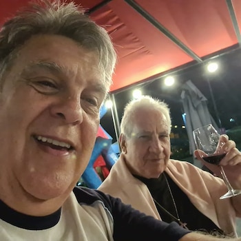 Selfie de Luis Ventura sonriendo en primer plano junto a Angelo Milano, quien sostiene una copa de vino, bajo una pérgola roja en un ambiente nocturno