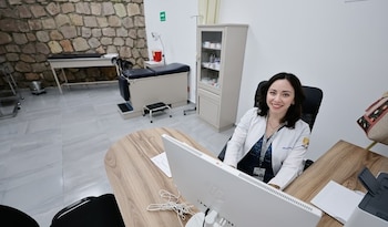 Una doctora sonriente en bata blanca sentada frente a un monitor de computadora en un consultorio médico, con una camilla y equipo médico detrás