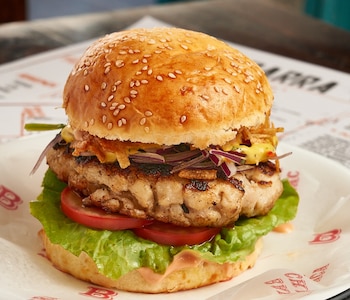 Primer plano de una hamburguesa con pan de sésamo, carne a la parrilla, lechuga, rodajas de tomate y cebolla morada, servida sobre papel de embalaje blanco y rojo