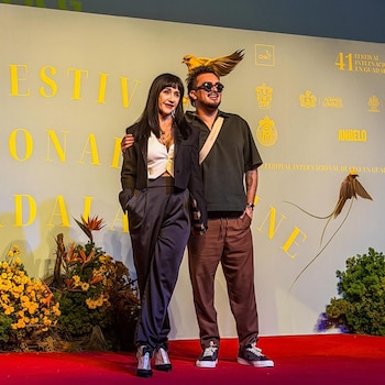 Una mujer de cabello oscuro y un hombre con gafas de sol posan de pie sobre una alfombra roja. Detrás, una pared clara con texto amarillo y logotipos