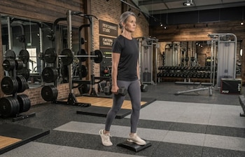 Mujer de cabello gris en un gimnasio, con mancuernas, un pie sobre una plataforma elevada, equipos de pesas, espejos y paredes de ladrillo