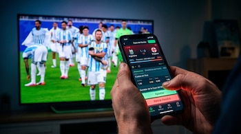 Unas manos sostienen un teléfono móvil con una aplicación de apuestas abierta. De fondo, una pantalla de televisión muestra a jugadores de fútbol en un campo verde.