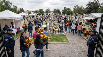 Cementerio con gente visitando tumbas adornadas con flores. Se ven carpas de información, puestos de venta y agentes municipales supervisando el acceso y tránsito.