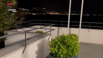 Vista nocturna desde un balcón con barandilla y vegetación, con el logo de Carabinieri Napoli en la esquina superior izquierda, y al fondo, luces de la ciudad lejana sobre el agua