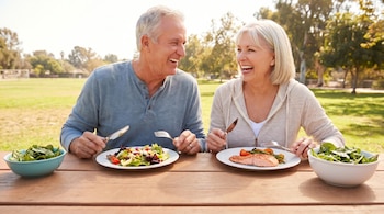 Pareja feliz come en mesa de picnic al aire libre. Hombre con ensalada, mujer con salmón y verduras. Bol de hojas verdes a la derecha.