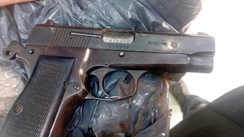 Primer plano de una pistola oscura, desgastada, con la inscripción 'DETECTIVE' y número de serie '415013', sobre una superficie de plástico negro arrugado