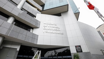 Fachada de edificio moderno con paneles grises y ventanas oscuras. Rótulo blanco "Ministerio de Vivienda, Construcción y Saneamiento". Número 3650. Bandera de Perú