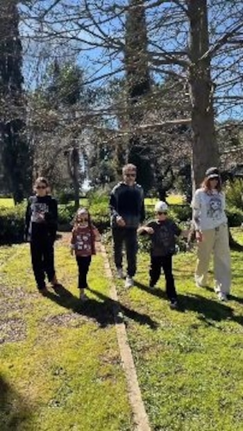 Cinco personas, dos adultos y tres niños, caminan sobre el césped de un jardín rodeado de árboles en un día soleado
