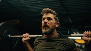 Primer plano de Aldo Kane, un hombre caucásico con barba, sosteniendo una barra con pesas sobre sus hombros, con el fondo oscuro de un gimnasio