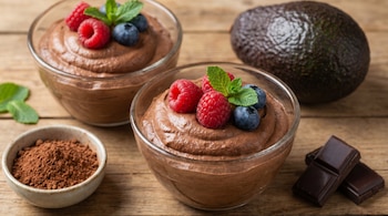 Mousse de chocolate en dos cuencos, decorado con frambuesas, arándanos y menta sobre una mesa de madera. Un aguacate, cacao en polvo y barras de chocolate se ven al lado.