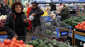 Personas comprando en un supermercado, con una mujer de pie junto a un carrito de compras y otra seleccionando productos de una gran exhibición de tomates y pepinos