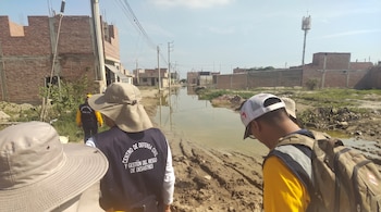 Personas con chalecos de Defensa Civil observan un camino de tierra inundado y embarrado entre viviendas de ladrillo