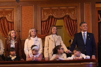 Un hombre y tres mujeres junto a tres niños en un palco de teatro con cortinas rojas y adornos dorados, observando un evento formal