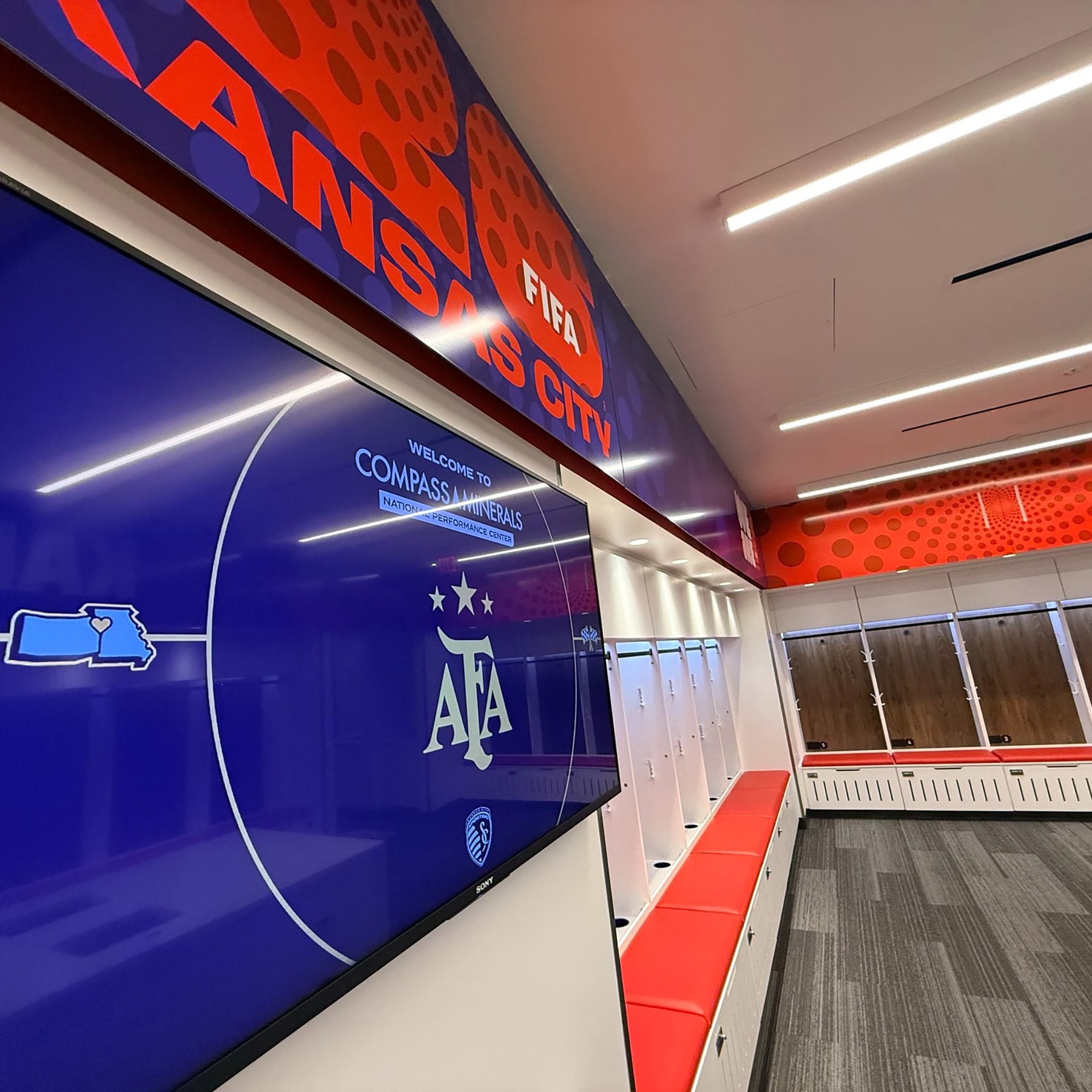 Un vestuario moderno en el Centro de Rendimiento Nacional Compass Minerals en Kansas City, con pantallas y mobiliario contemporáneo, mostrando el logo de la AFA (@afaseleccion)