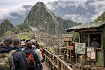 Una fila de personas en una pasarela de madera, un puesto de tickets, carteles informativos, la ciudadela de Machu Picchu, montañas y neblina.