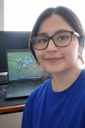 Una mujer joven con cabello oscuro y gafas de montura negra sonríe a la cámara. Detrás, un portátil abierto muestra una imagen de ovejas en una pradera