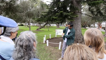 Hombre con chaqueta verde muestra un folleto a un grupo de personas en un parque con césped, árboles y un cerco blanco