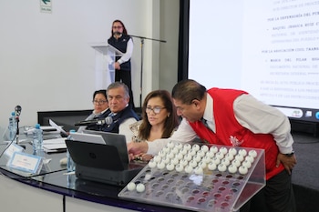 Sala con cuatro personas, dos en mesa con laptop y bandeja de bolas numeradas, una en atril y otra sentada. Pantalla con texto y botellas