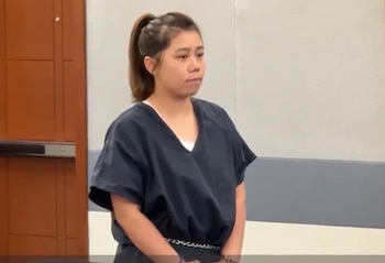 Mujer joven de cabello oscuro recogido en una coleta, vestida con uniforme de reclusa oscuro, con esposas en las muñecas, de pie