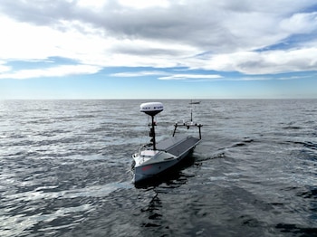 Un vehículo de superficie no tripulado (USV) Lightfish gris navega en aguas oscuras, bajo un cielo azul parcialmente nublado, con otro USV más pequeño al fondo