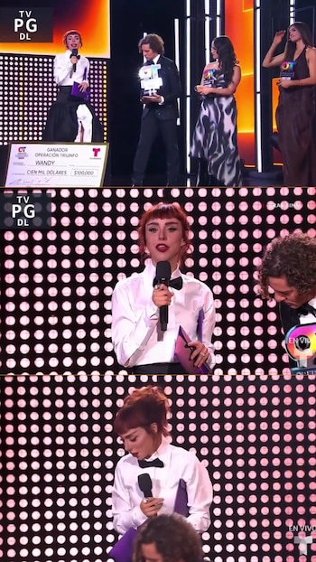 Mujer pelirroja con micrófono y pajarita, hombre con trofeo, cheque de cien mil dólares para Wandy, dos mujeres de fondo, panel de luces LED