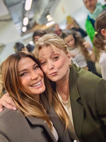 Dos mujeres posan en primer plano para una fotografía. Una de cabello castaño sonríe y la otra de cabello rubio frunce los labios. Hay personas detrás